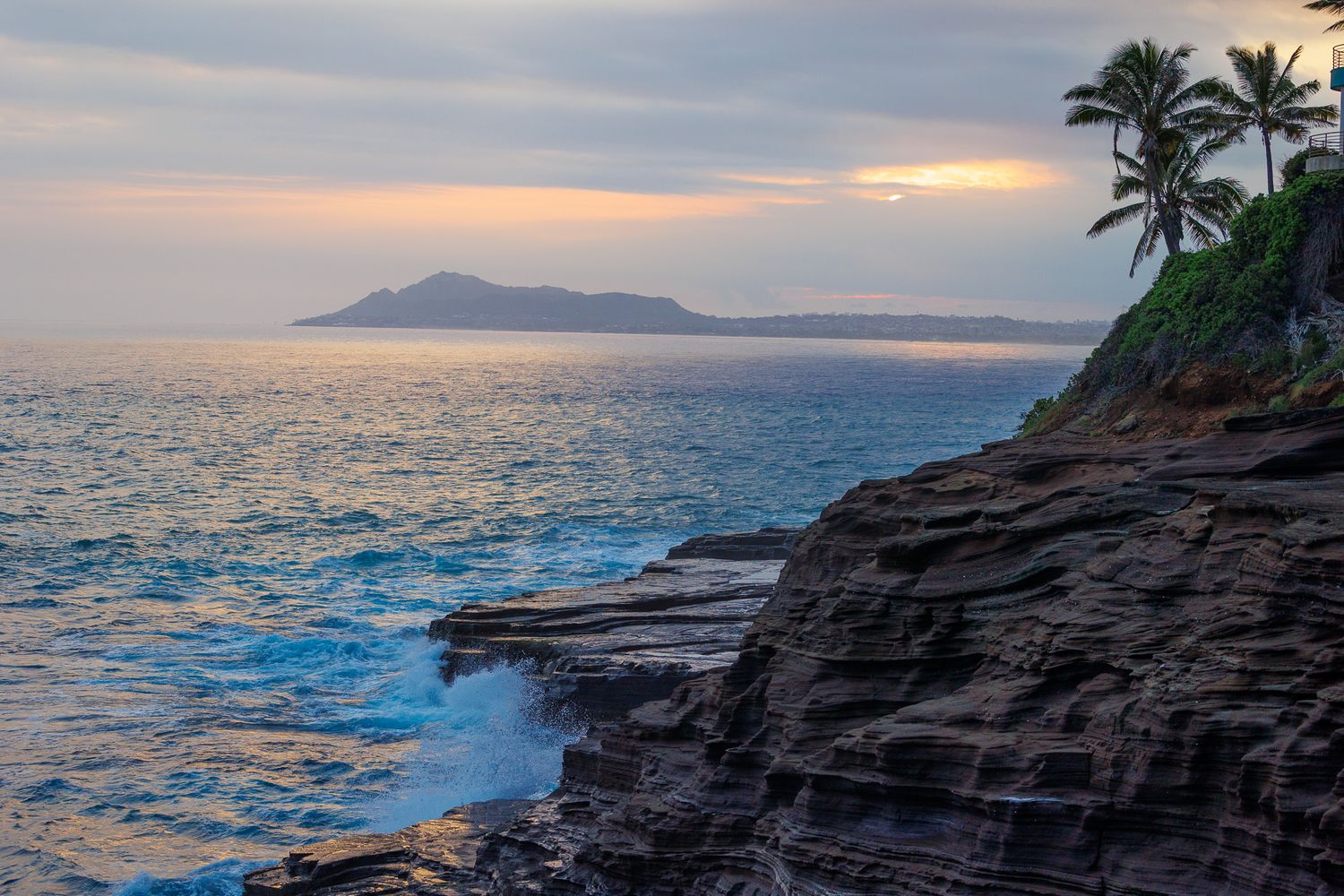 Dramatic sunset over rocky Hawaiian coastline with palm trees and distant island on horizon.