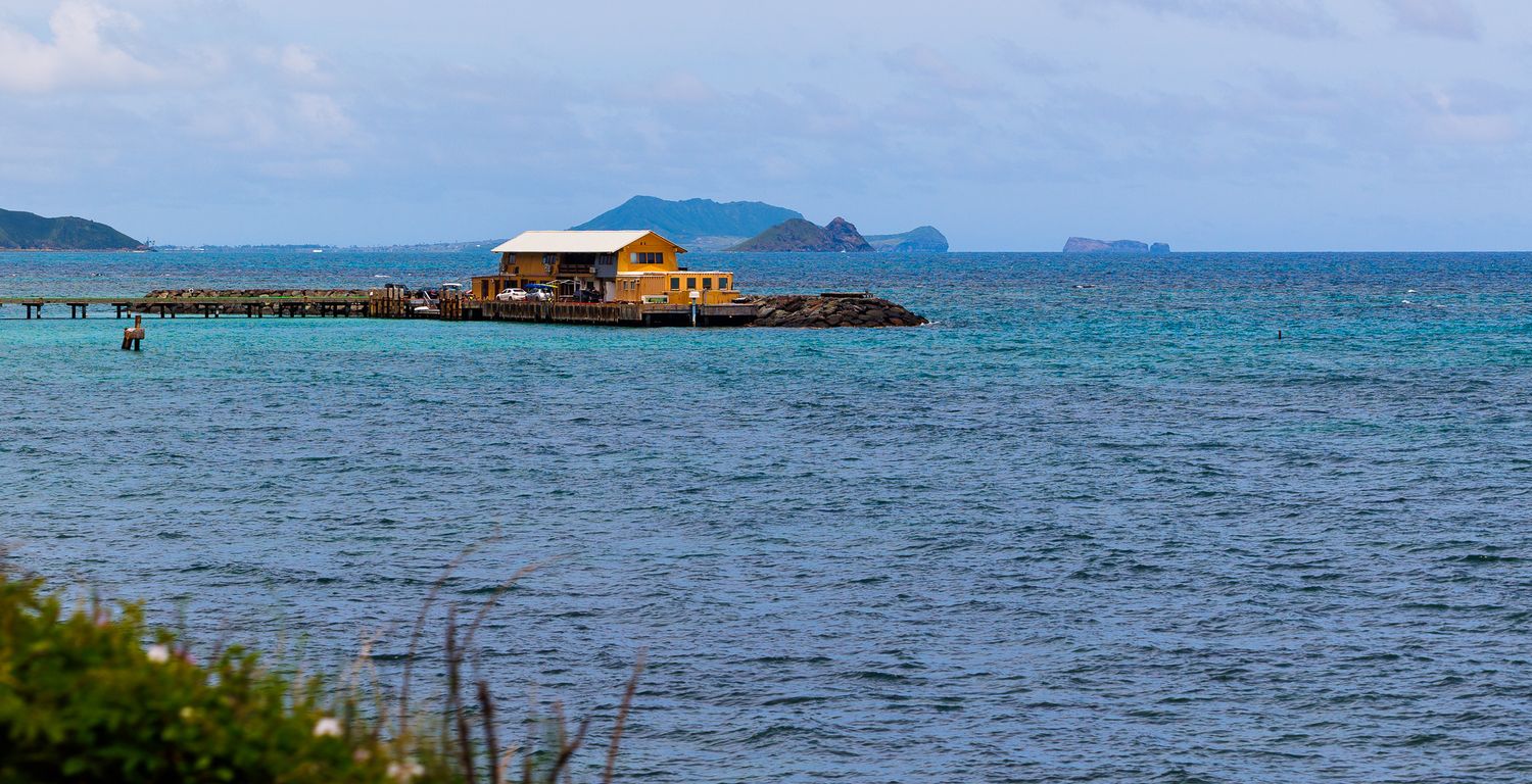 Tropical overwater bungalow on a pier surrounded by turquoise sea with distant islands under a partly cloudy sky.
