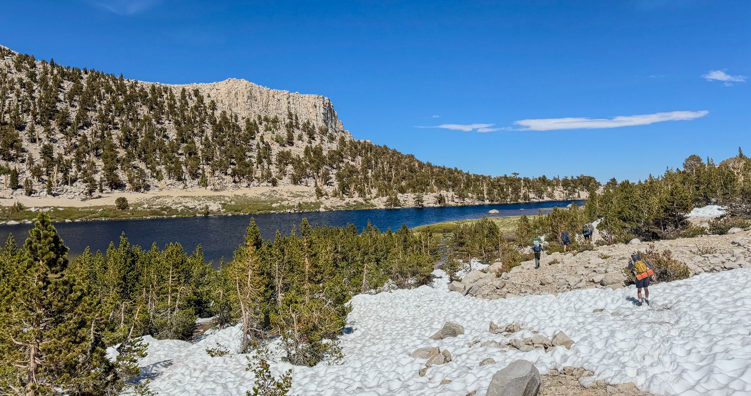 Hiker exploring a snowy alpine lake trail surrounded by pine trees and rocky cliffs under a clear blue sky.