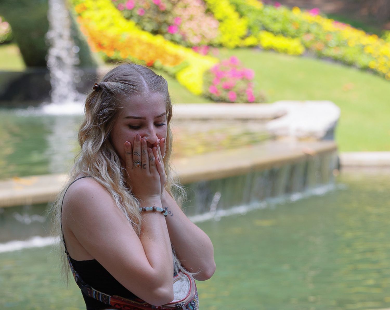 Surprised blonde woman covering her mouth with hands near a beautiful garden fountain.