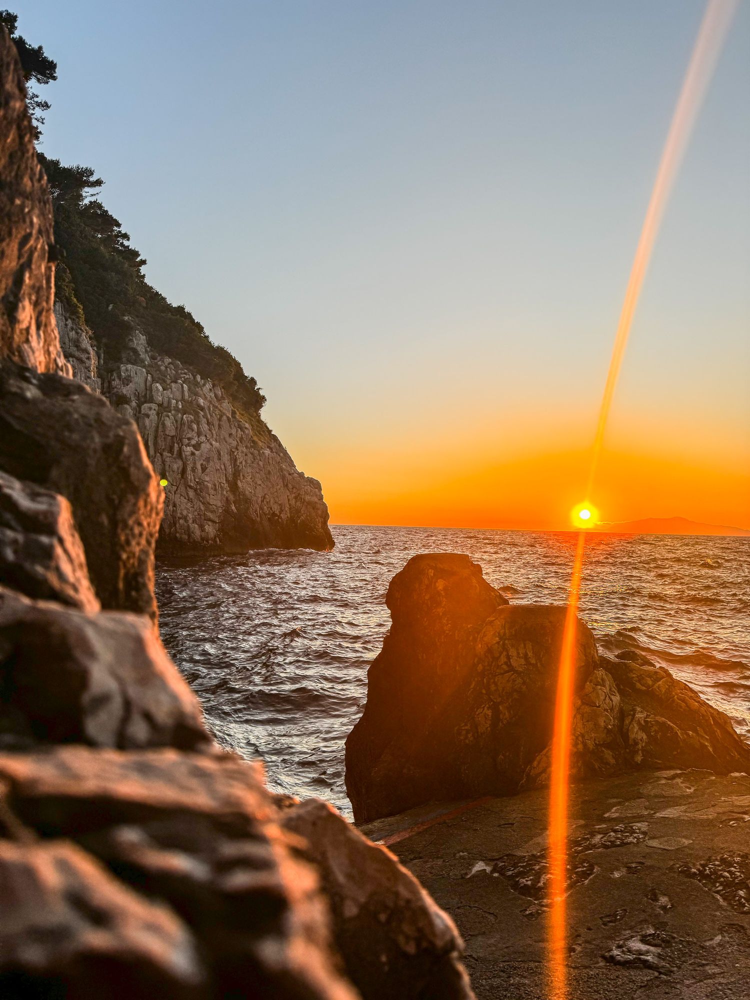 Stunning golden sunset over rocky Mediterranean coastline with dramatic sun flare and cliff views.