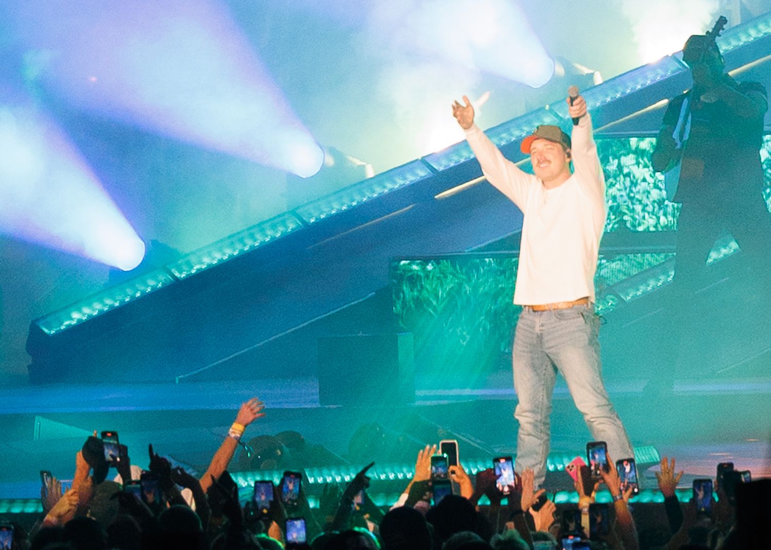Male performer raises arms triumphantly on stage bathed in teal and green concert lighting.