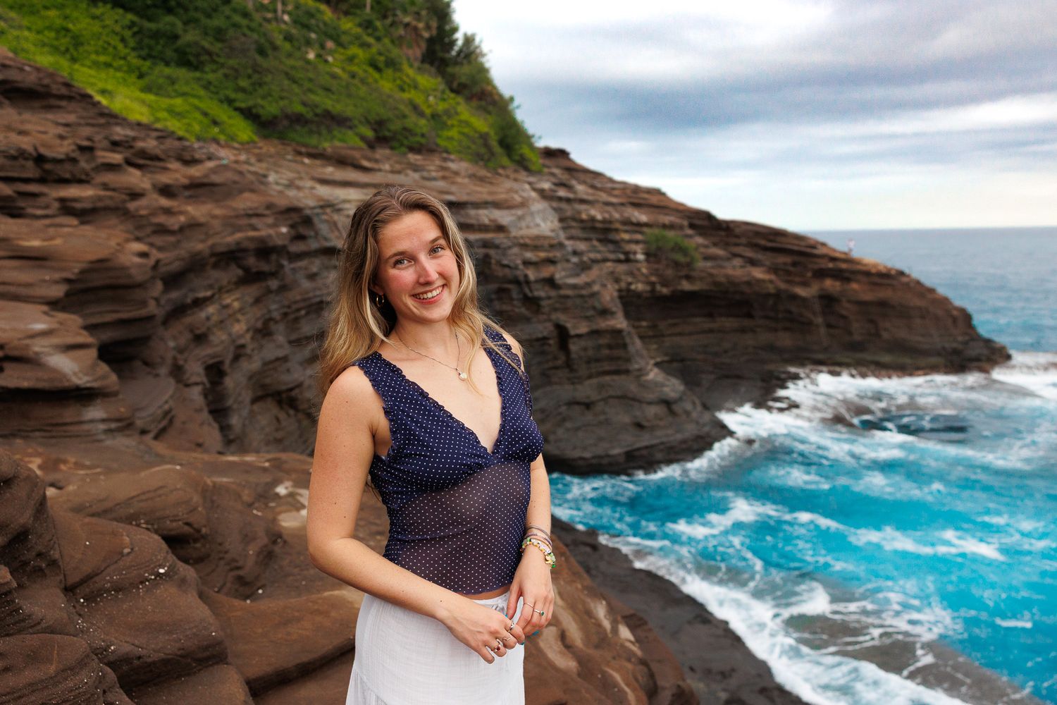 Young woman smiling on rocky Hawaiian coastline with turquoise waves and layered red cliffs under cloudy skies.