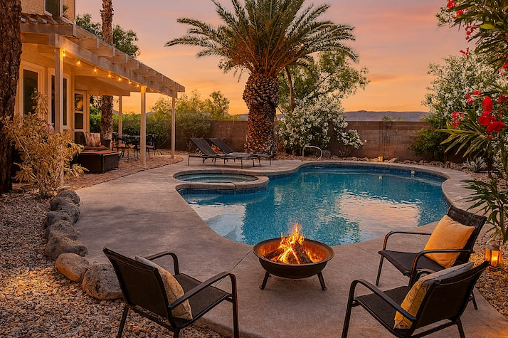 Backyard oasis at dusk with a fire pit, pool, spa, palm trees, and string lights on a covered patio.