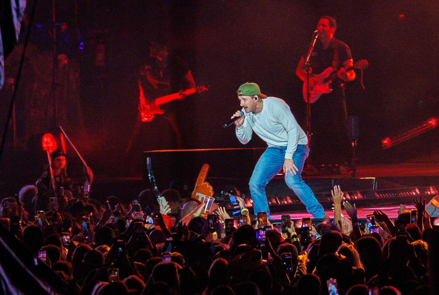 Male singer performs energetically on stage before large crowd with band and red guitar.