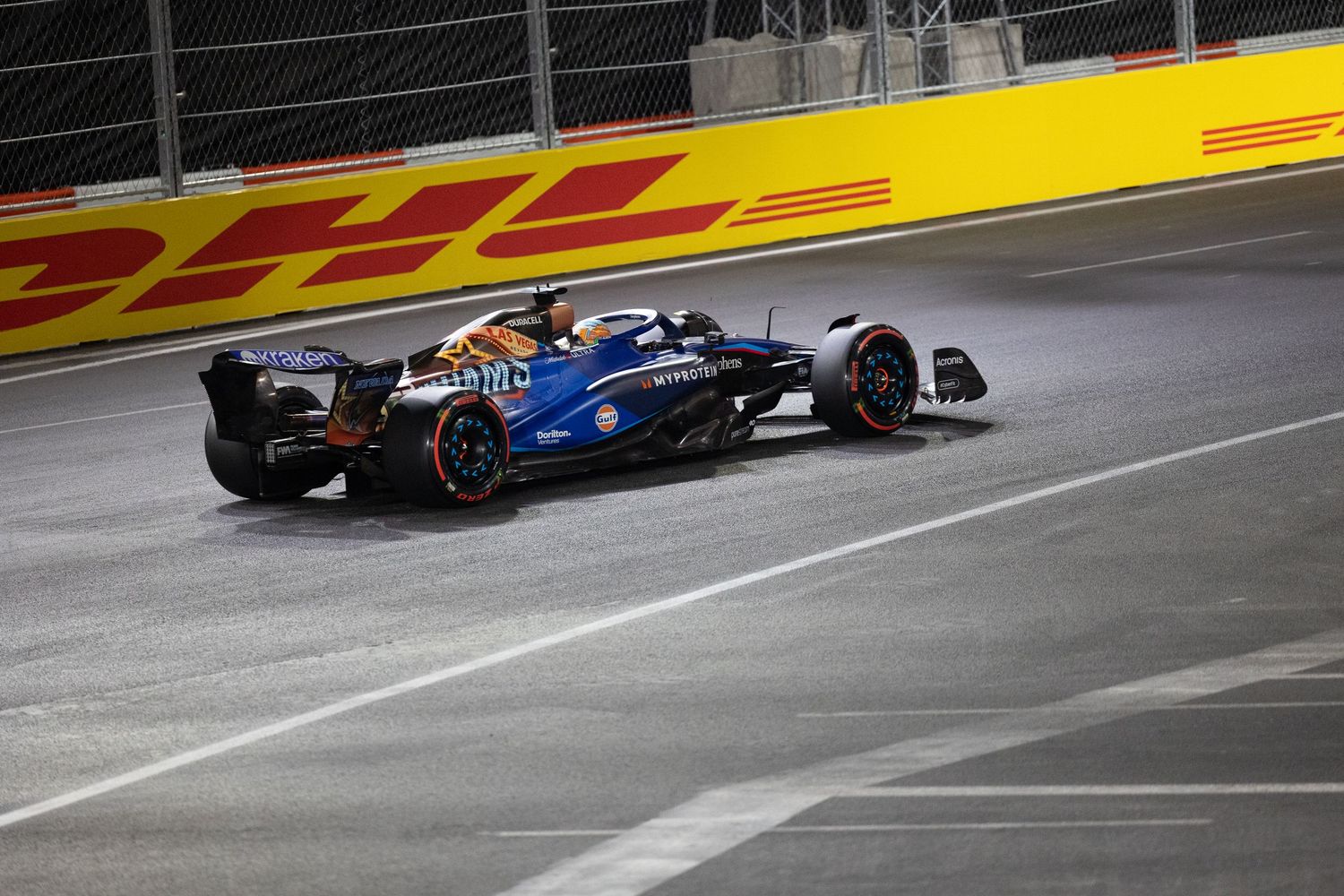 Blue Williams F1 racing car speeding on track at night race with DHL advertising boards.