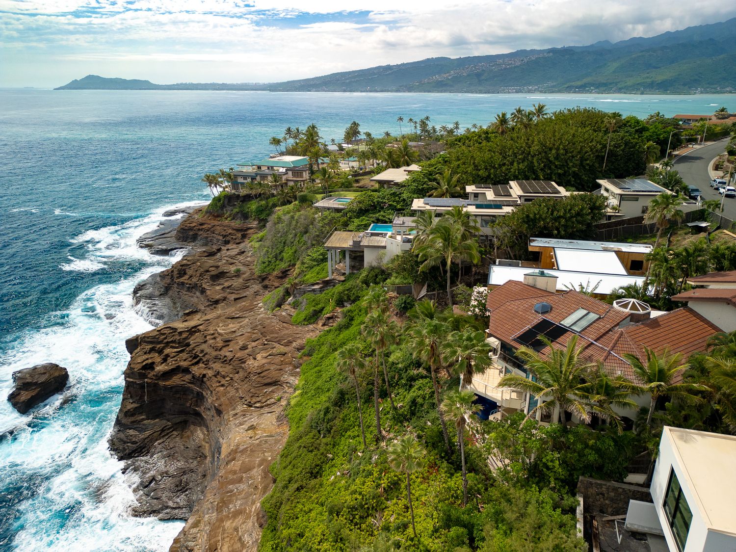 Aerial view of luxury cliffside homes overlooking rocky coastline with turquoise ocean waves in Hawaii.