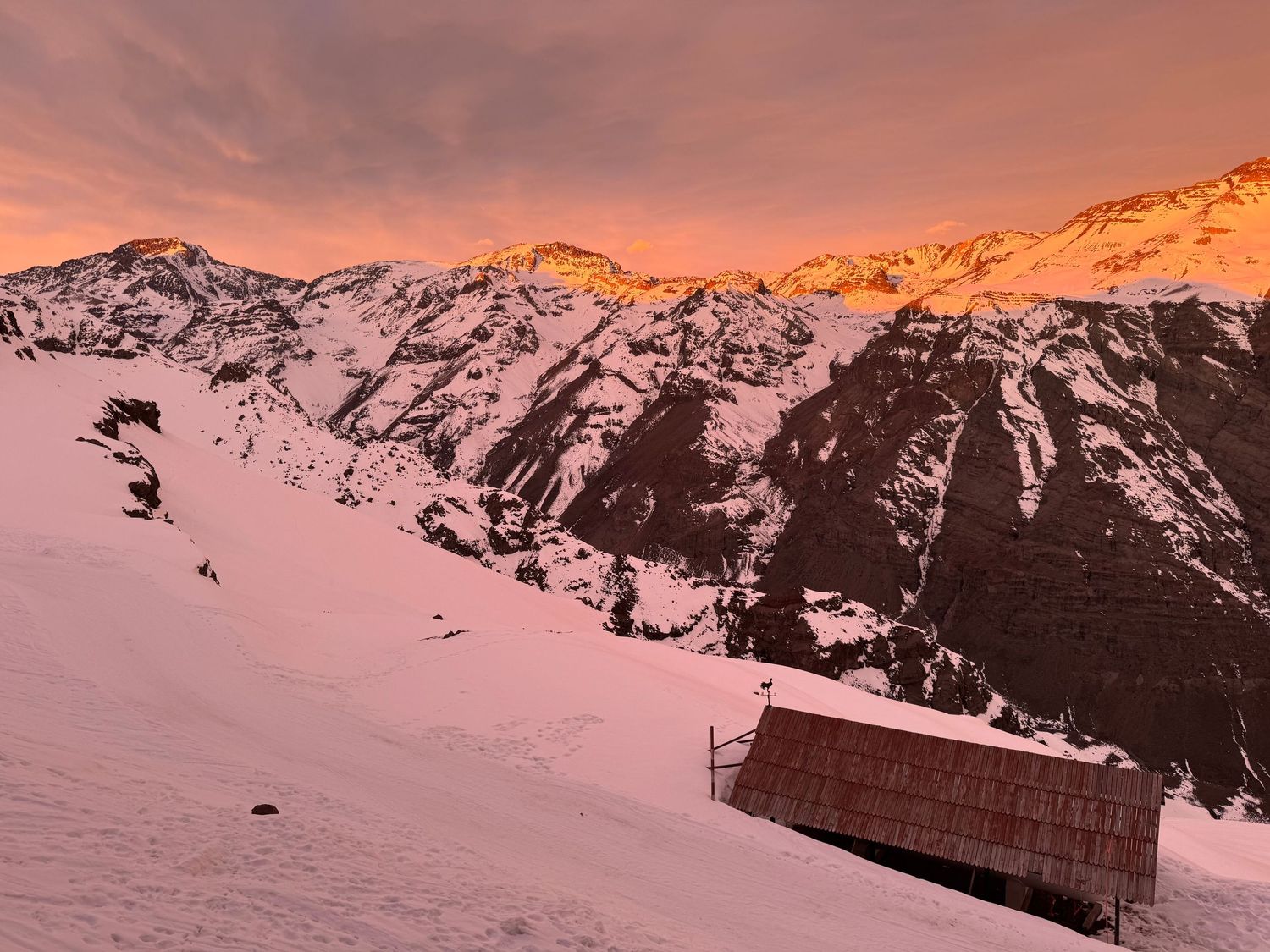 Dramatic alpine sunset casts orange glow over snow-covered mountain peaks with small shelter.