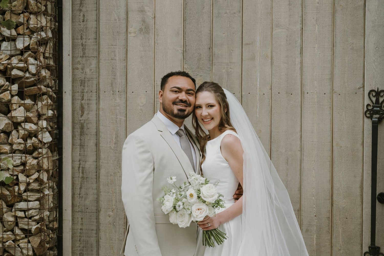 Smiling bride and groom pose together in front of rustic wooden doors, bride holding white floral bouquet.