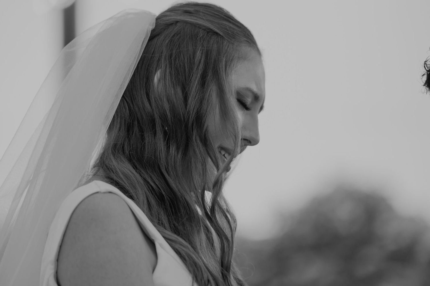 A beautiful bride wearing a white dress and veil looks down in a serene black and white wedding portrait.