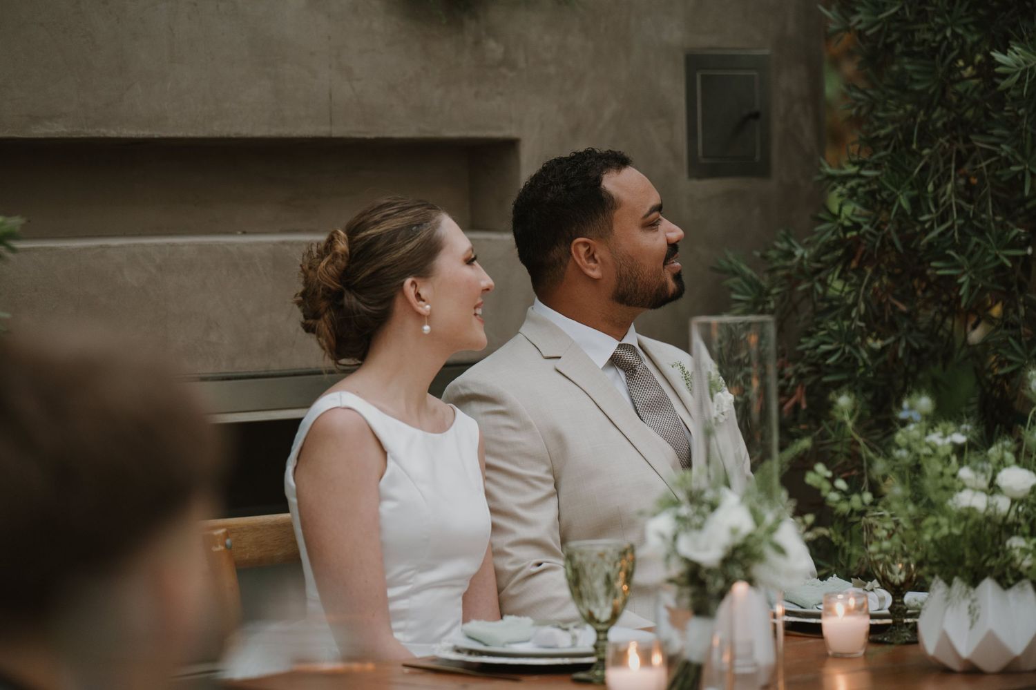 Bride and groom seated at wedding reception table with candles and greenery decor in romantic setting.