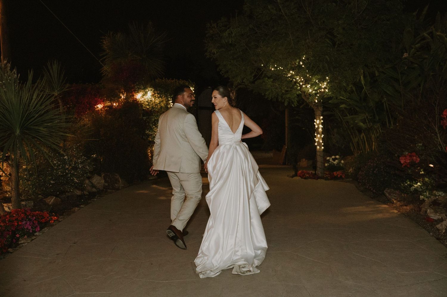 Bride and groom sharing romantic moment on garden path surrounded by twinkling lights at night.