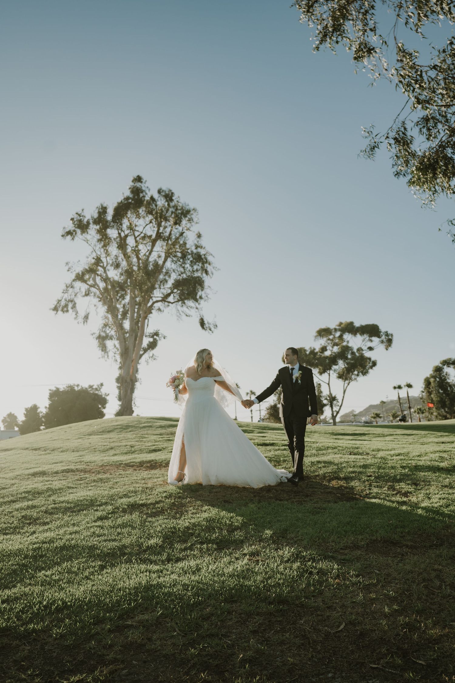 Bride and groom walking hand in hand across a sunlit green hill on their wedding day.