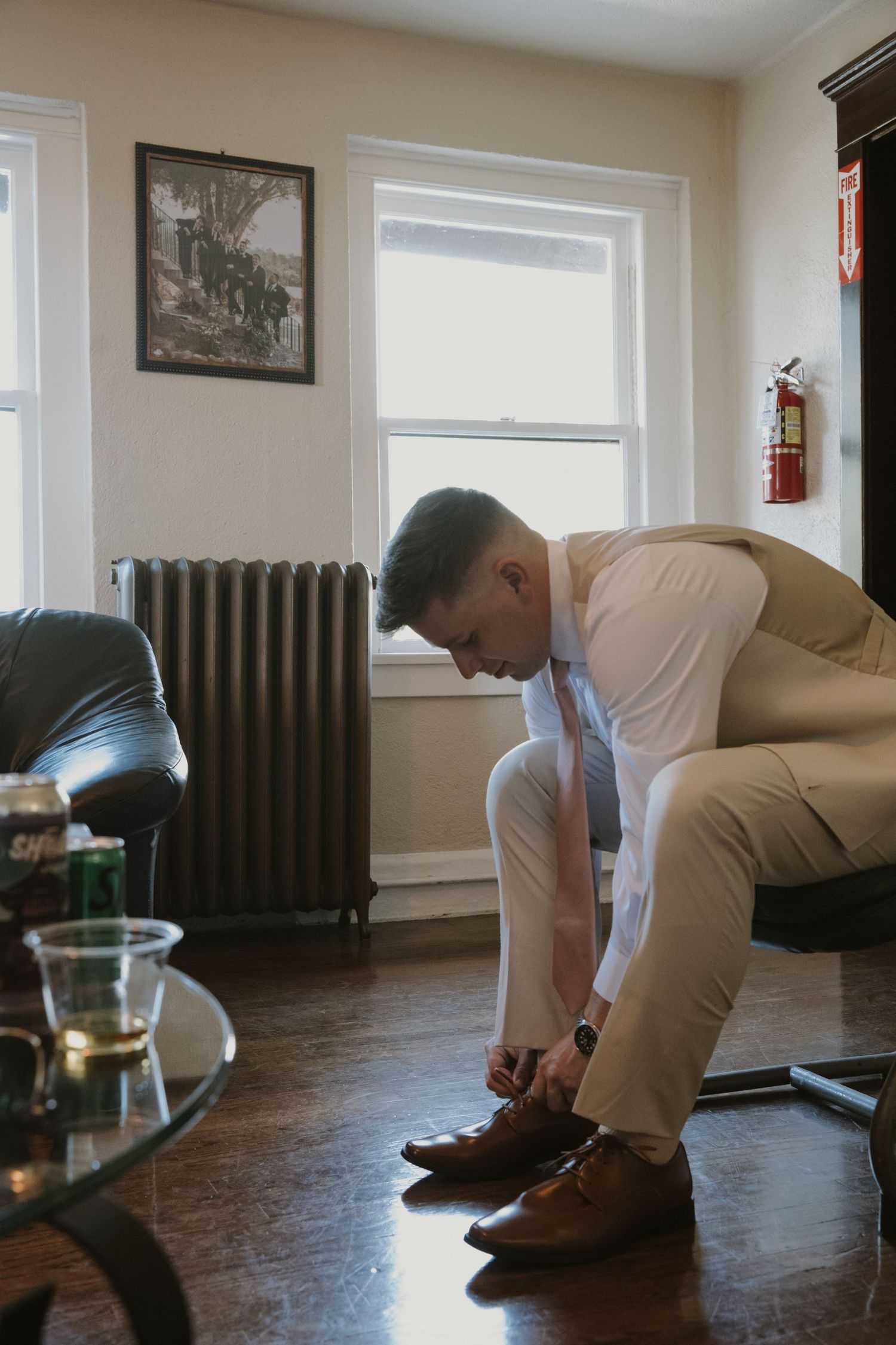 Groom in tan suit tying brown dress shoes before wedding ceremony in vintage room with radiator.