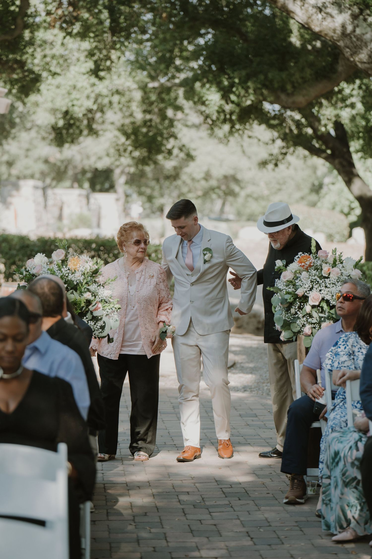 Groom in white suit escorts elderly woman down brick wedding aisle flanked by floral arrangements.