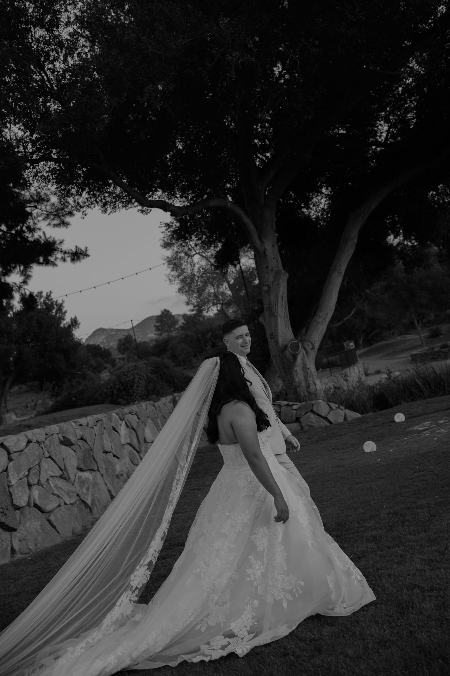 Bride in wedding gown with long lace veil flowing in black and white photo outdoors near stone wall and large tree.
