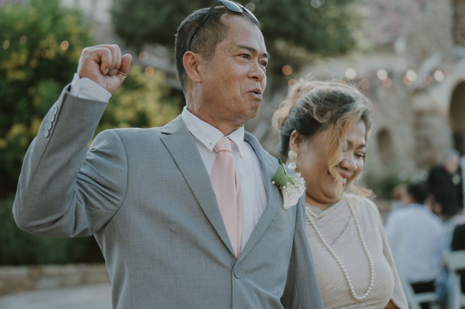 Proud father in gray suit raises fist celebrating at outdoor wedding ceremony.