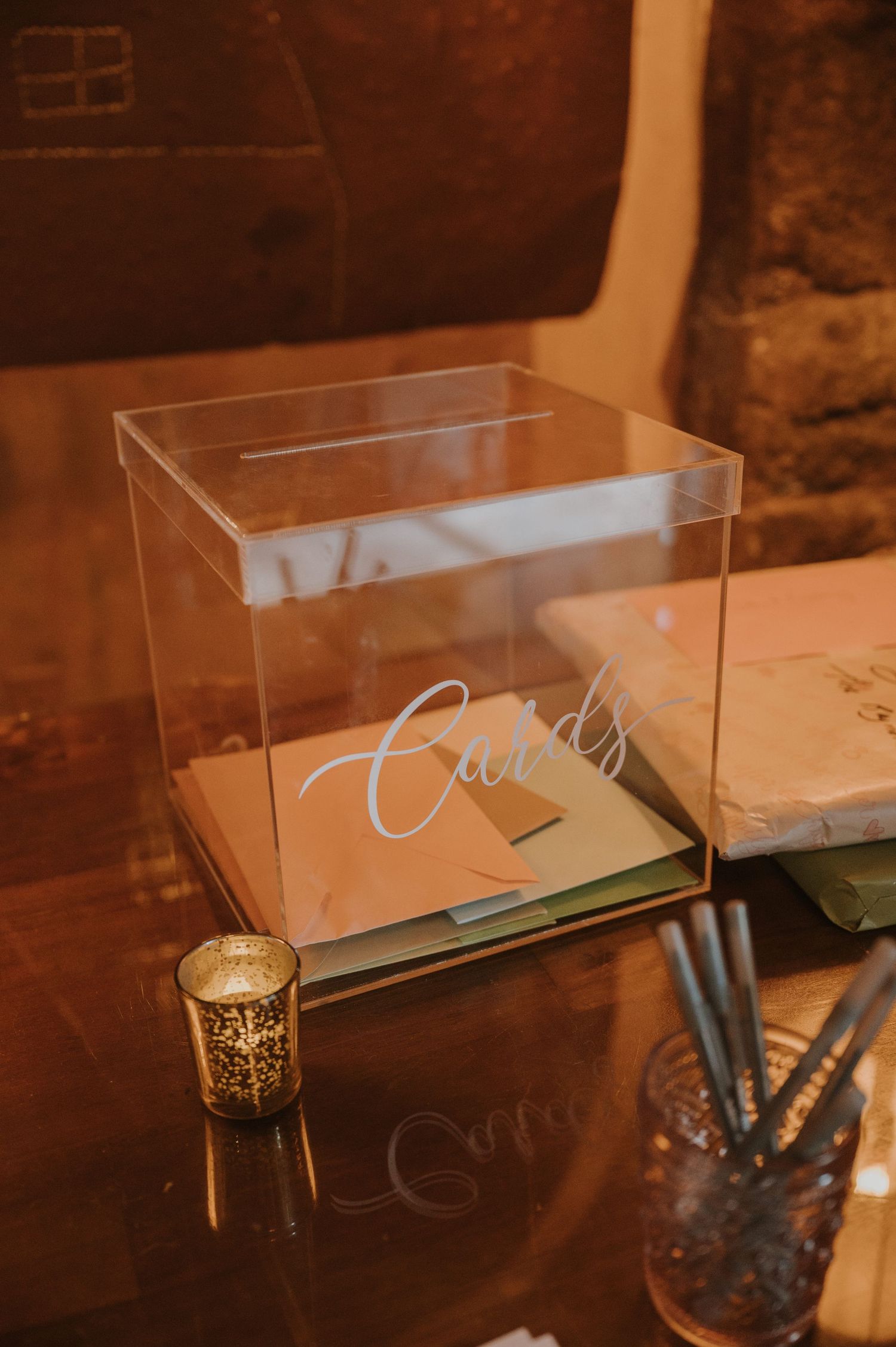 Clear acrylic wedding card box with cursive Cards label displayed on a reflective table with gold votive candle.