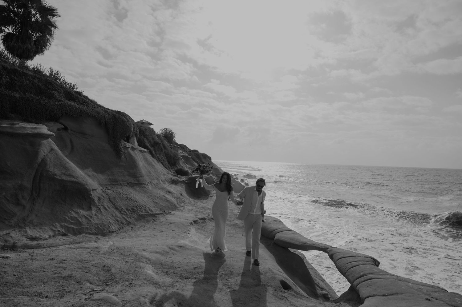 Black and white photo of couple walking on sandy beach path beside rocky cliffs and ocean waves.