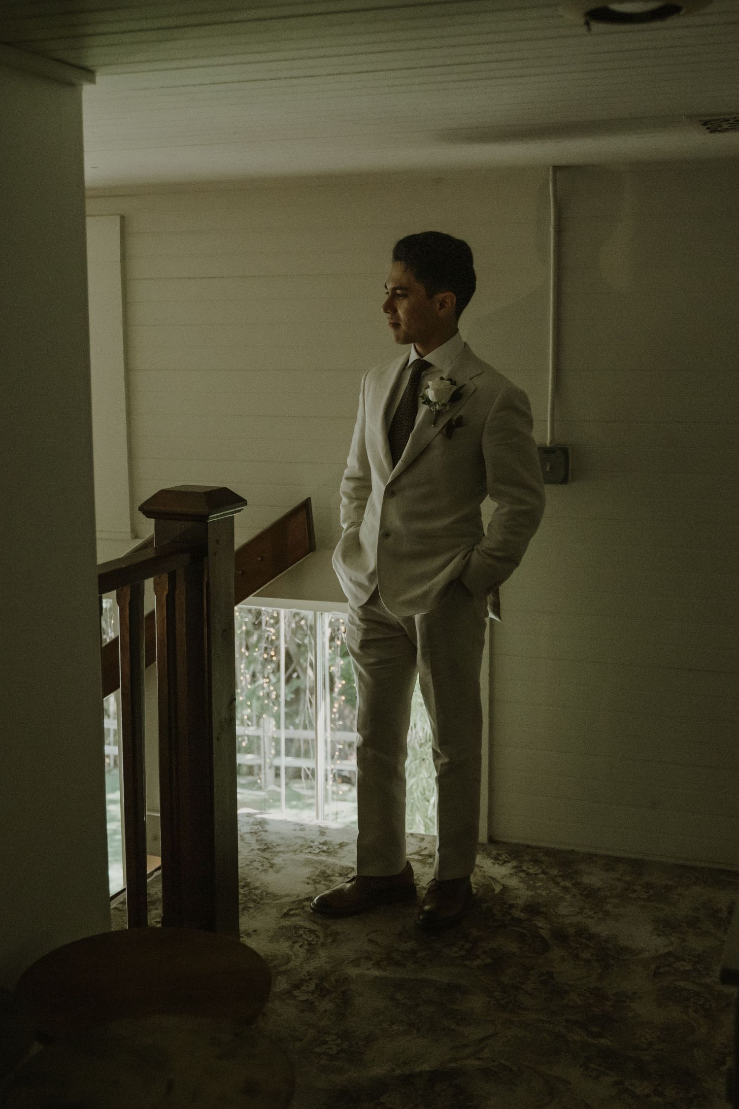 Groom in white suit with boutonniere stands by staircase railing, gazing toward window in dimly lit room.