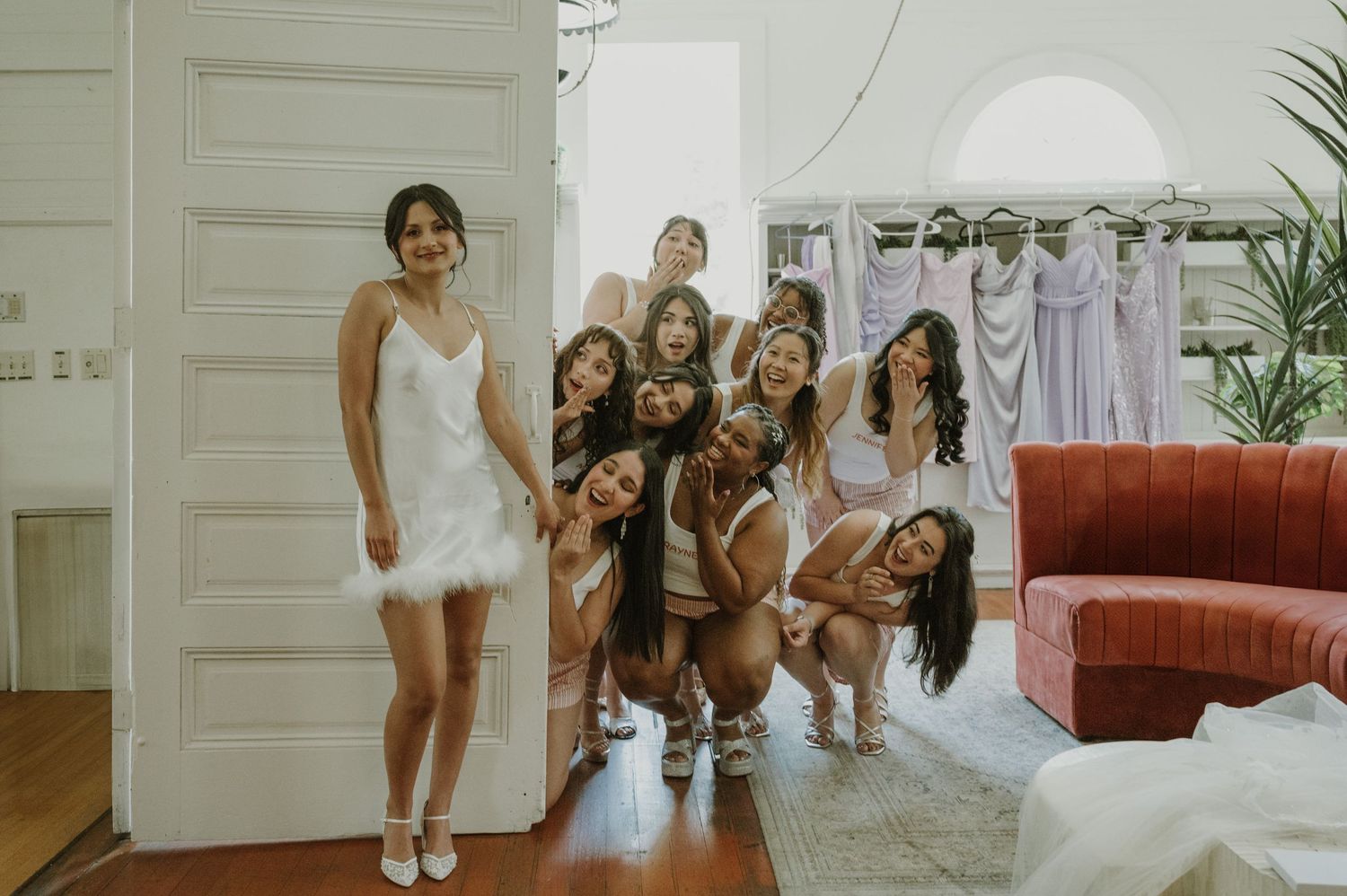 Excited bridesmaids posing playfully with bride in white feather dress by an open door.