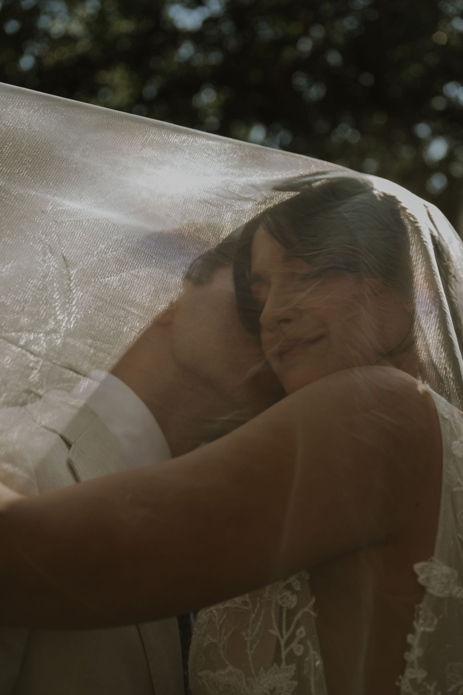 Bride and groom share an intimate embrace under a sheer lace wedding veil outdoors.