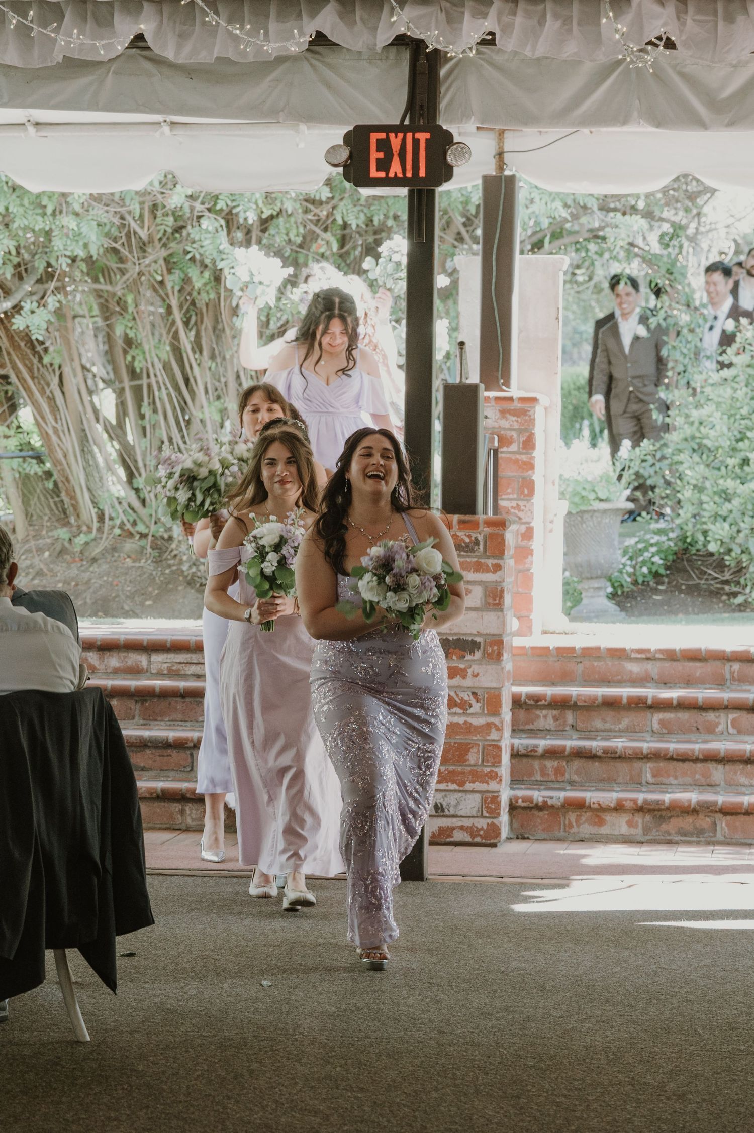Bridesmaids in sequin and pastel gowns carrying bouquets walk through a brick-framed venue entrance.
