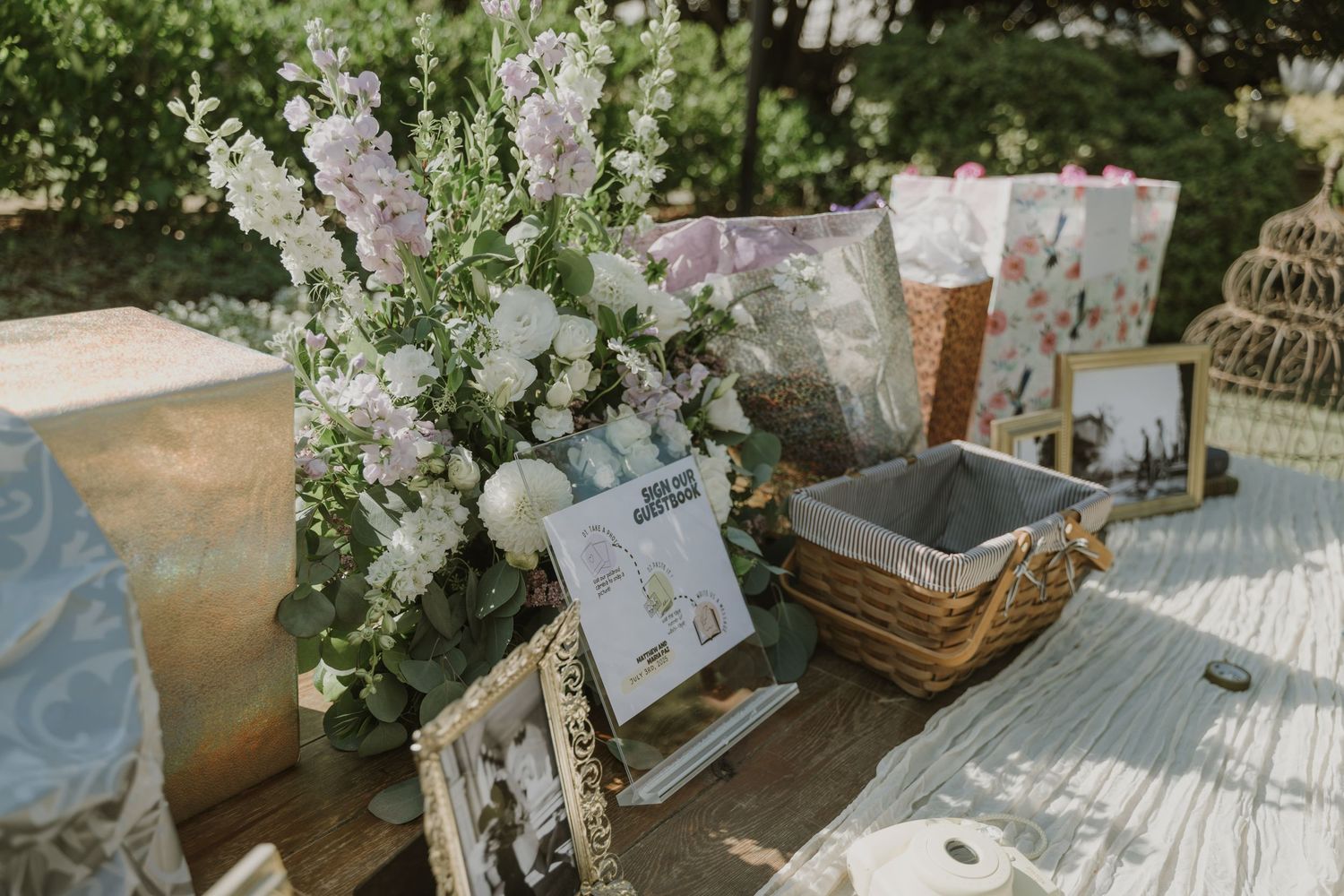 Elegant wedding gift table with floral arrangements, framed photos, wicker basket, and decorative birdcage outdoors.