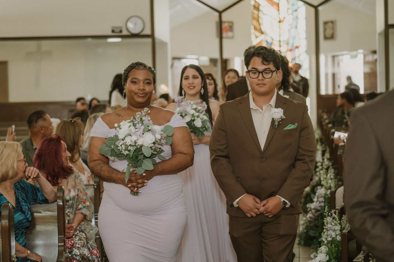 Bridesmaid in white gown walks down church aisle with groomsman in brown suit during wedding ceremony.