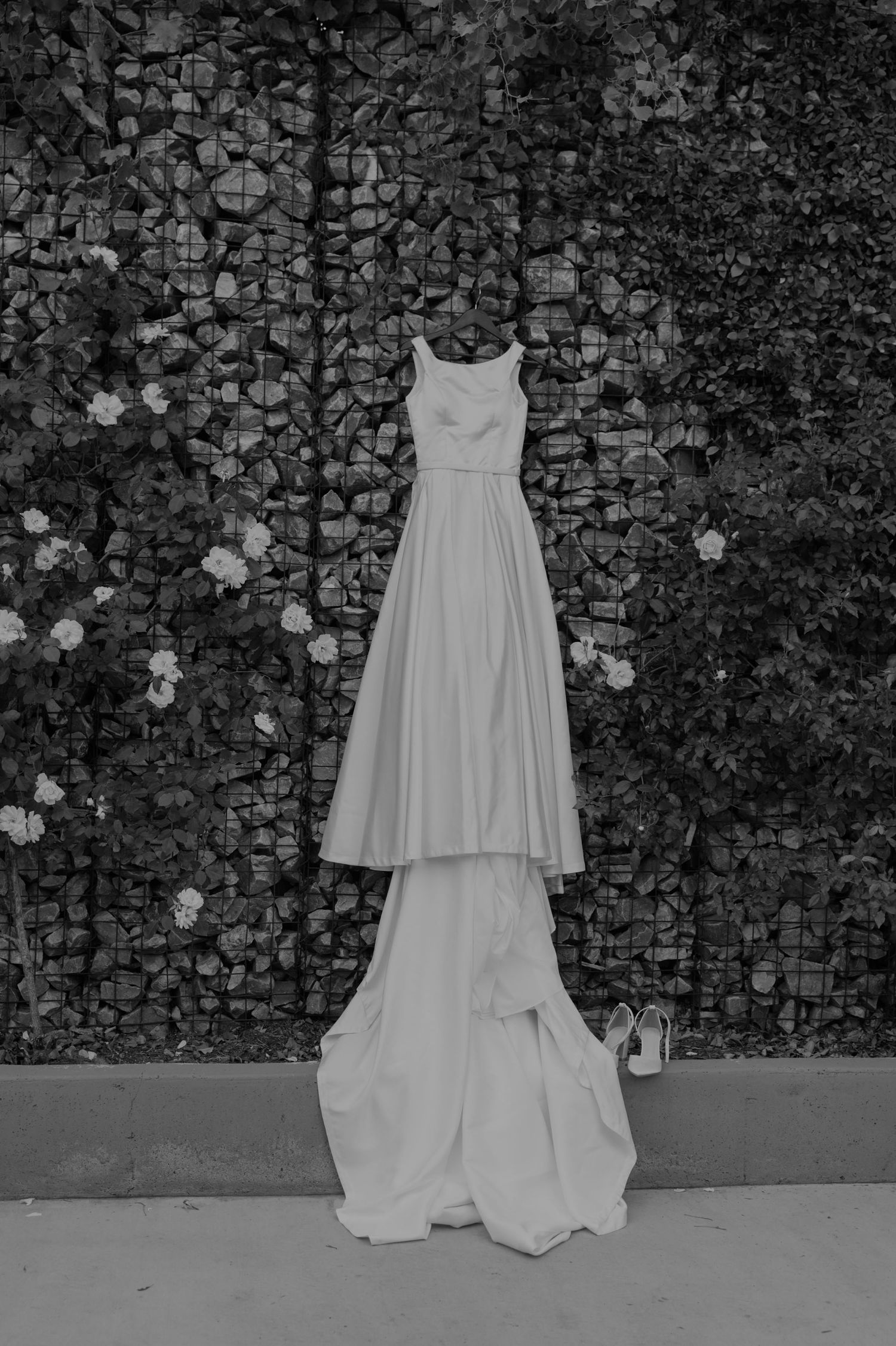 Elegant sleeveless wedding gown with long train hanging outdoors against stone wall with white flowers.