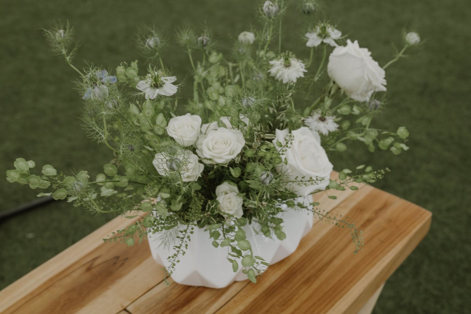 White roses and greenery arranged in a white geometric vase on a wooden table at an outdoor wedding.