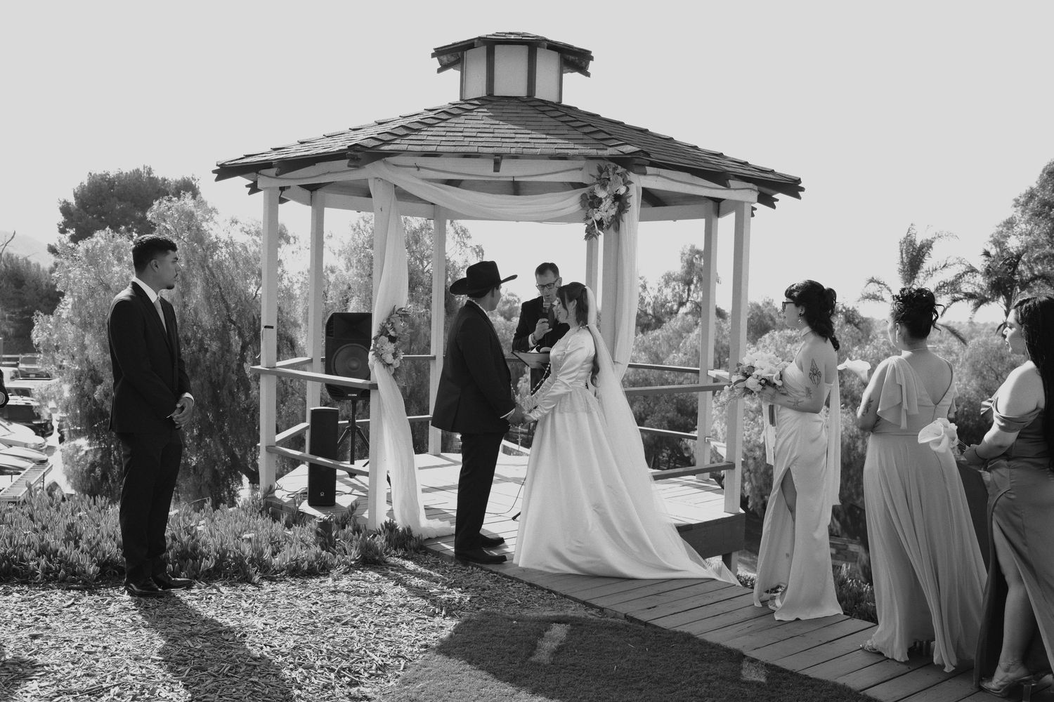 Black and white outdoor wedding ceremony at a wooden gazebo with bride, groom, officiant, and guests.
