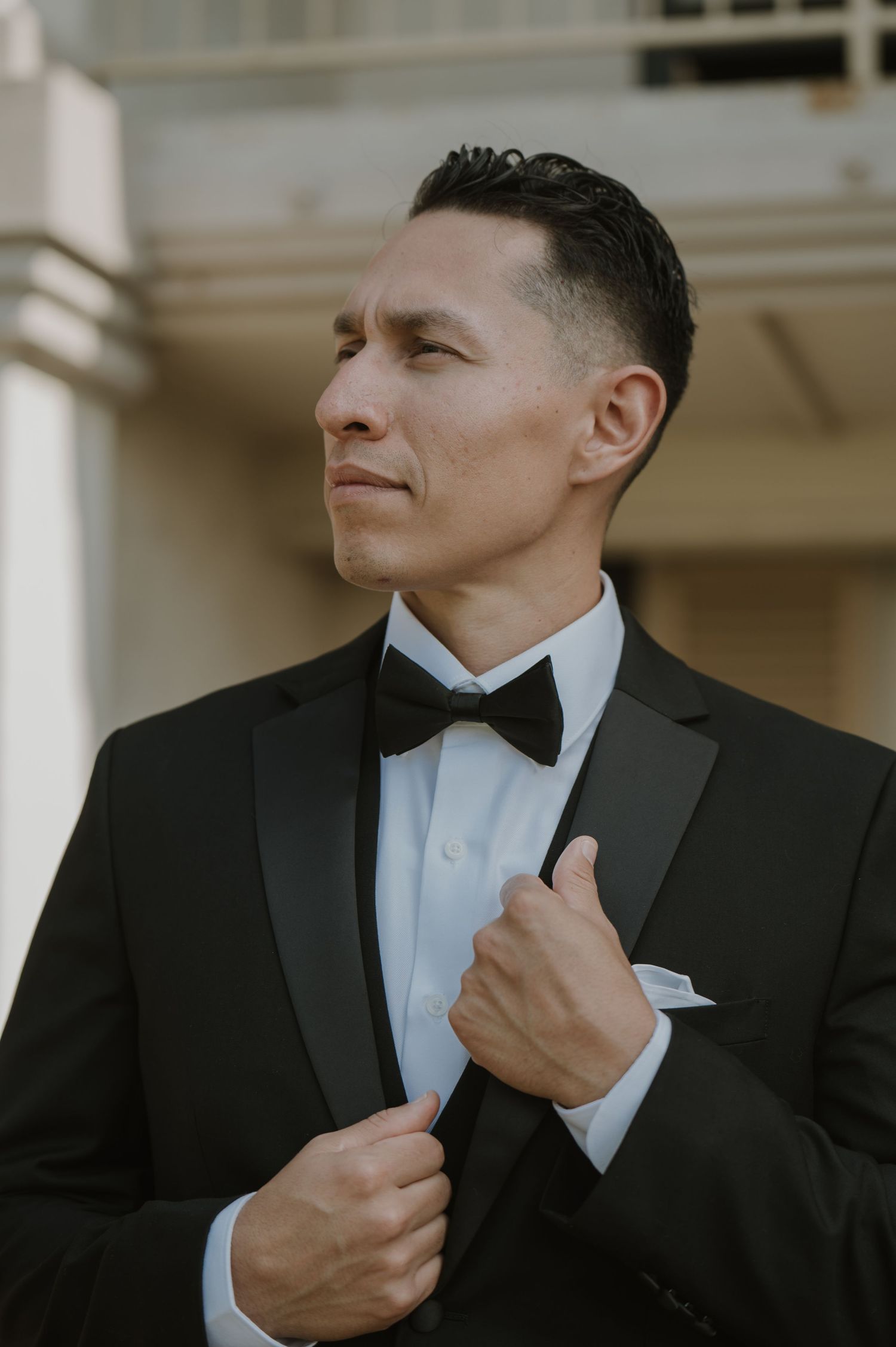 Groom in classic black tuxedo and bow tie adjusting lapel outdoors.