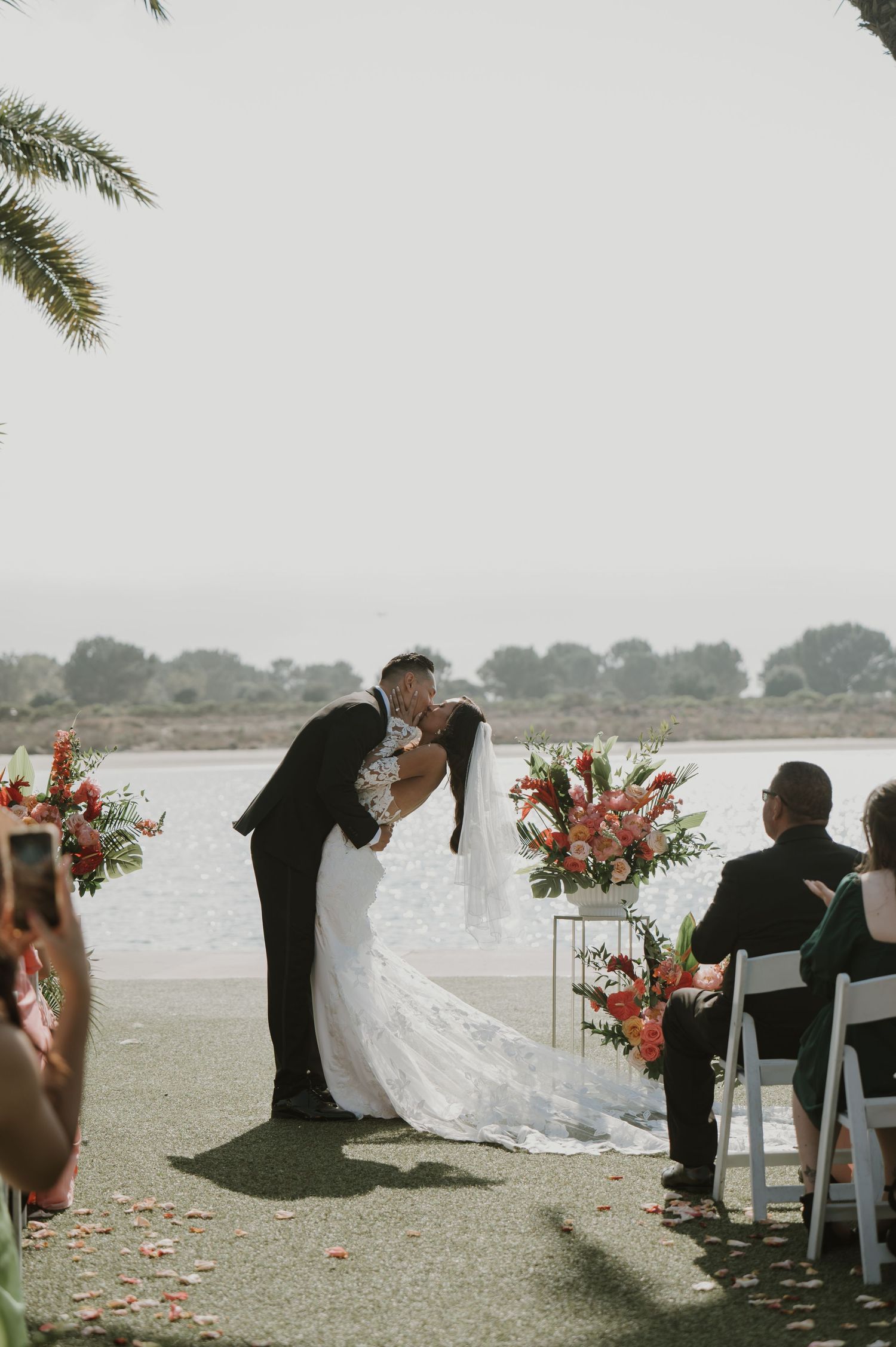 Romantic beach wedding ceremony setup with white chairs and coral floral arrangements by the water.