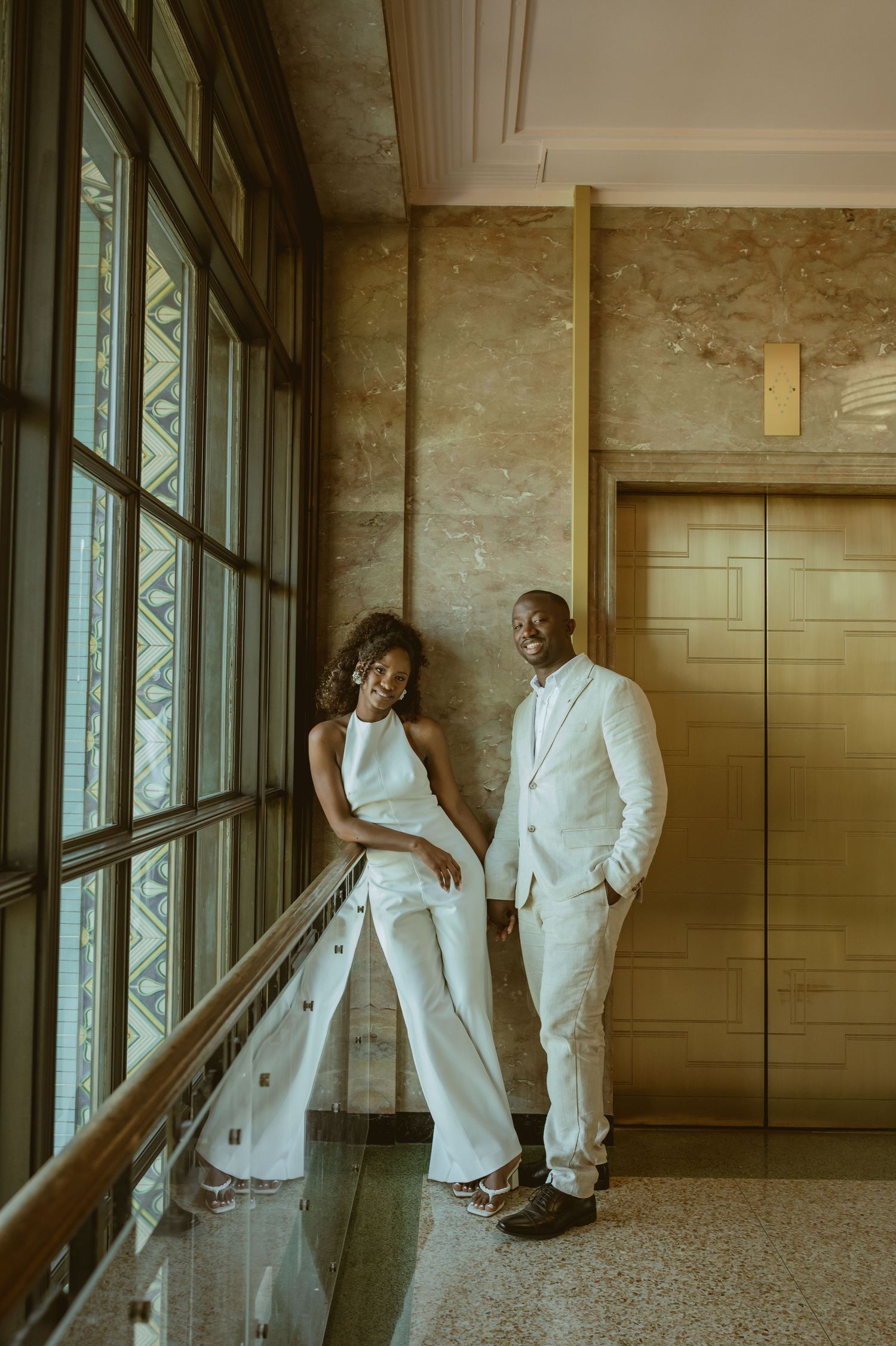 A couple in elegant white outfits sharing a romantic moment by large windows.