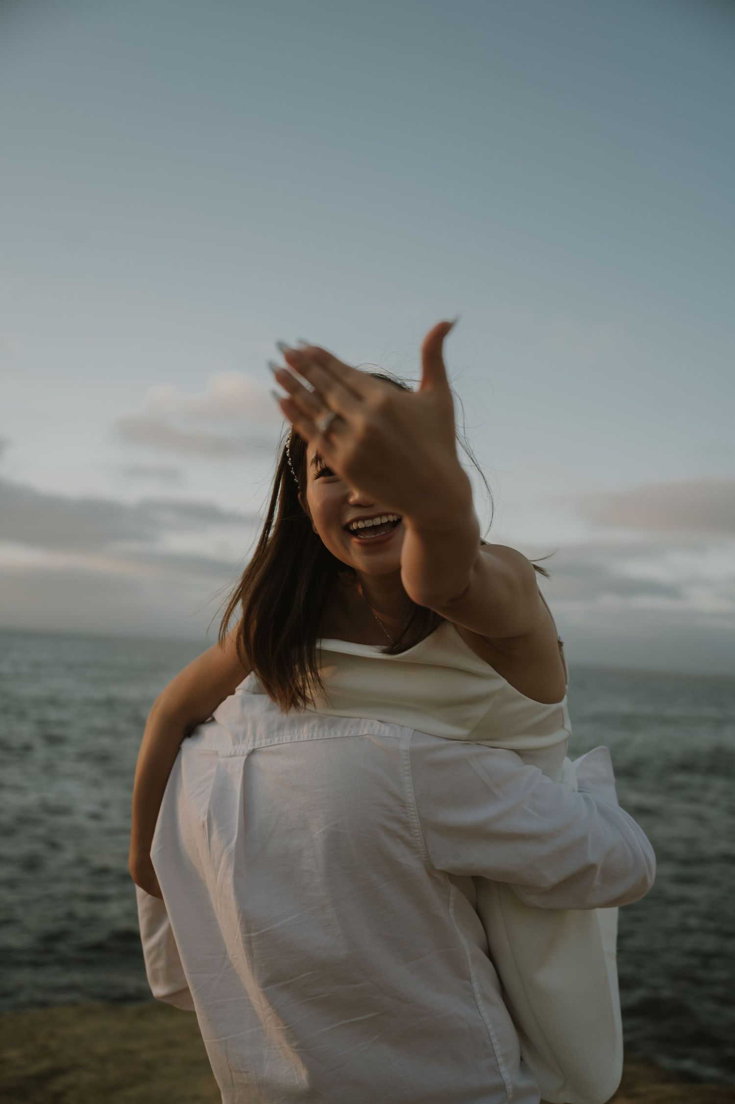 A couple in white clothing embraces joyfully against a moody ocean backdrop.