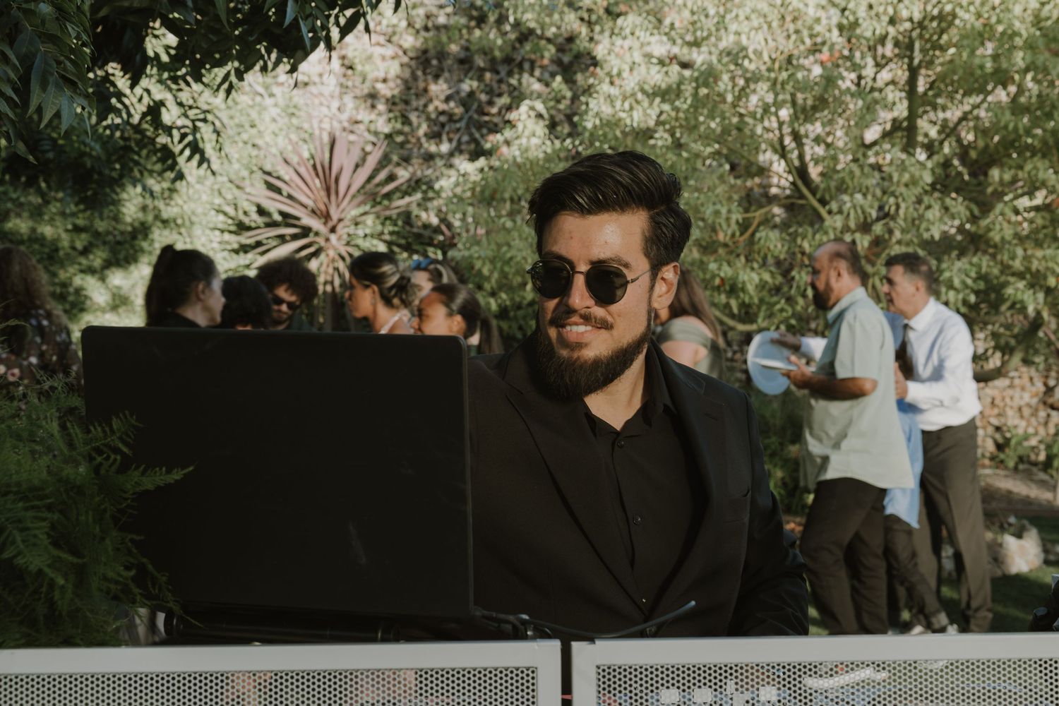 Smiling DJ in black suit and sunglasses performing at an outdoor wedding reception surrounded by greenery.
