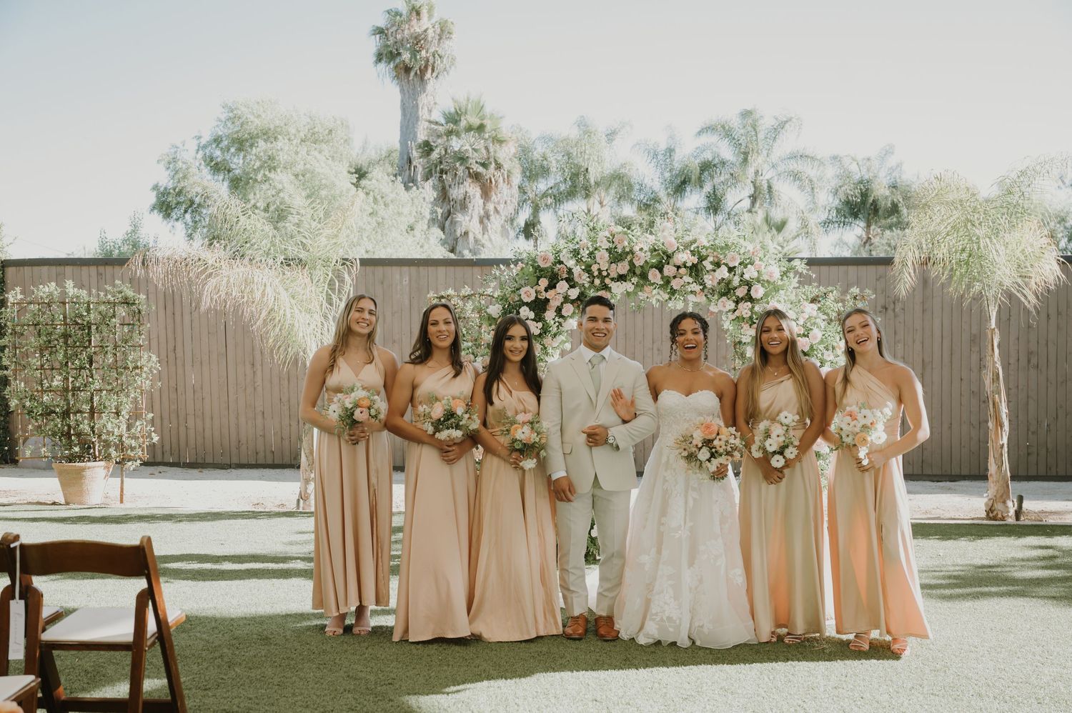 Bride in white lace gown poses with five bridesmaids in champagne dresses before floral arch outdoors.