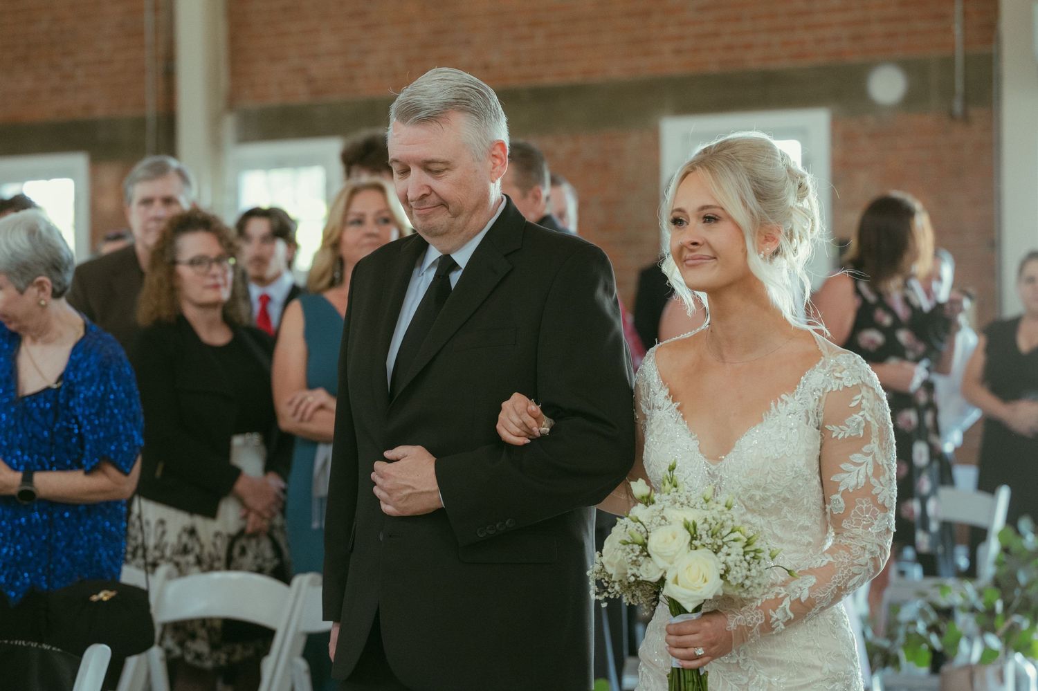 Father escorts smiling bride in lace wedding gown down the aisle at indoor brick wedding venue.