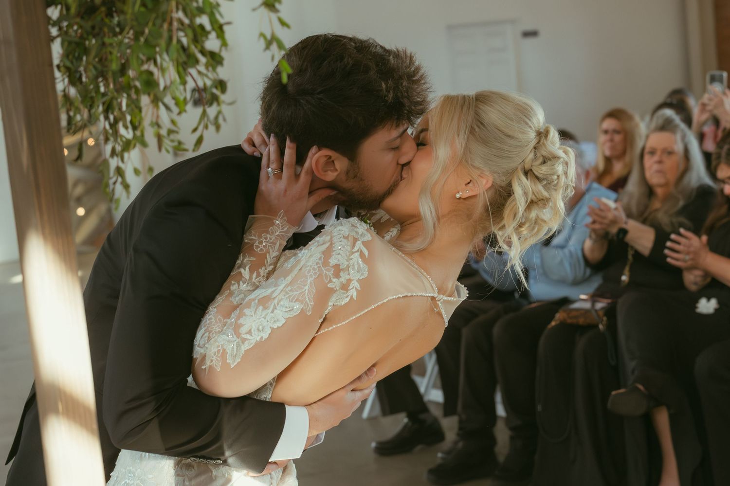 Bride and groom share their first kiss at wedding ceremony while guests applaud in elegant venue.