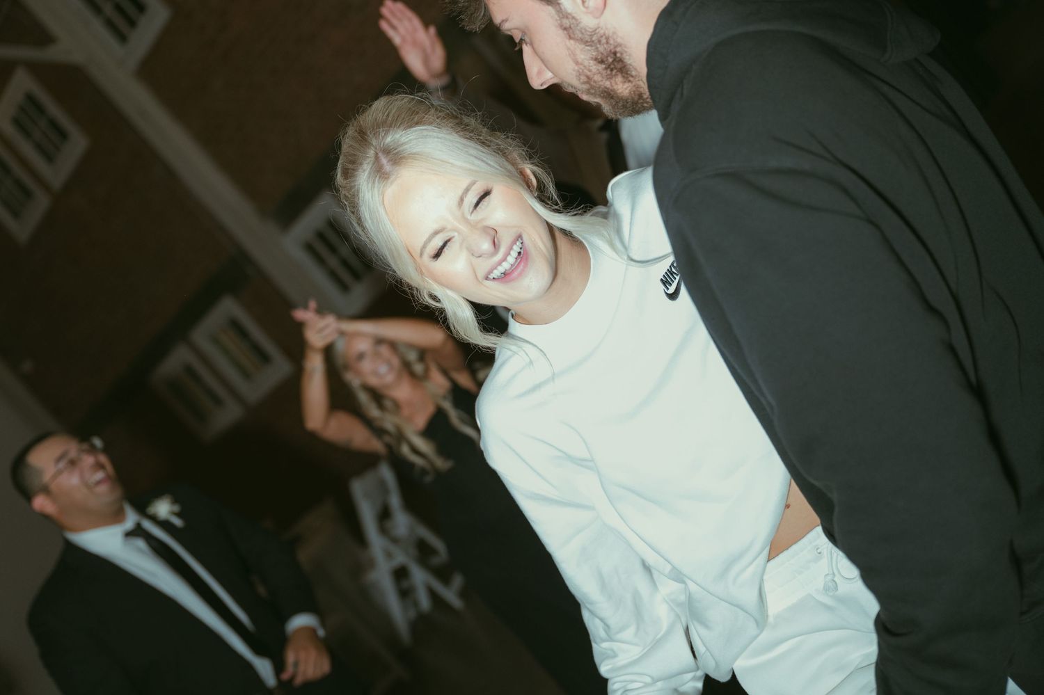 Bride laughing joyfully with groom during wedding reception while guests celebrate in background.