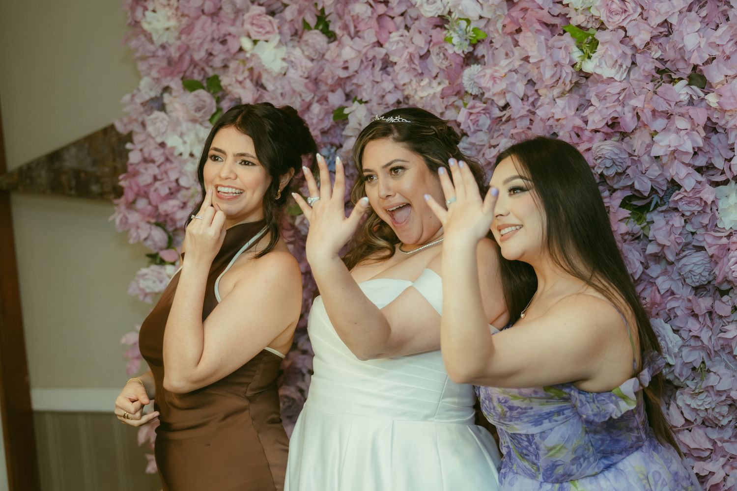 Bride poses joyfully with two bridesmaids in front of a stunning pink floral wall backdrop.