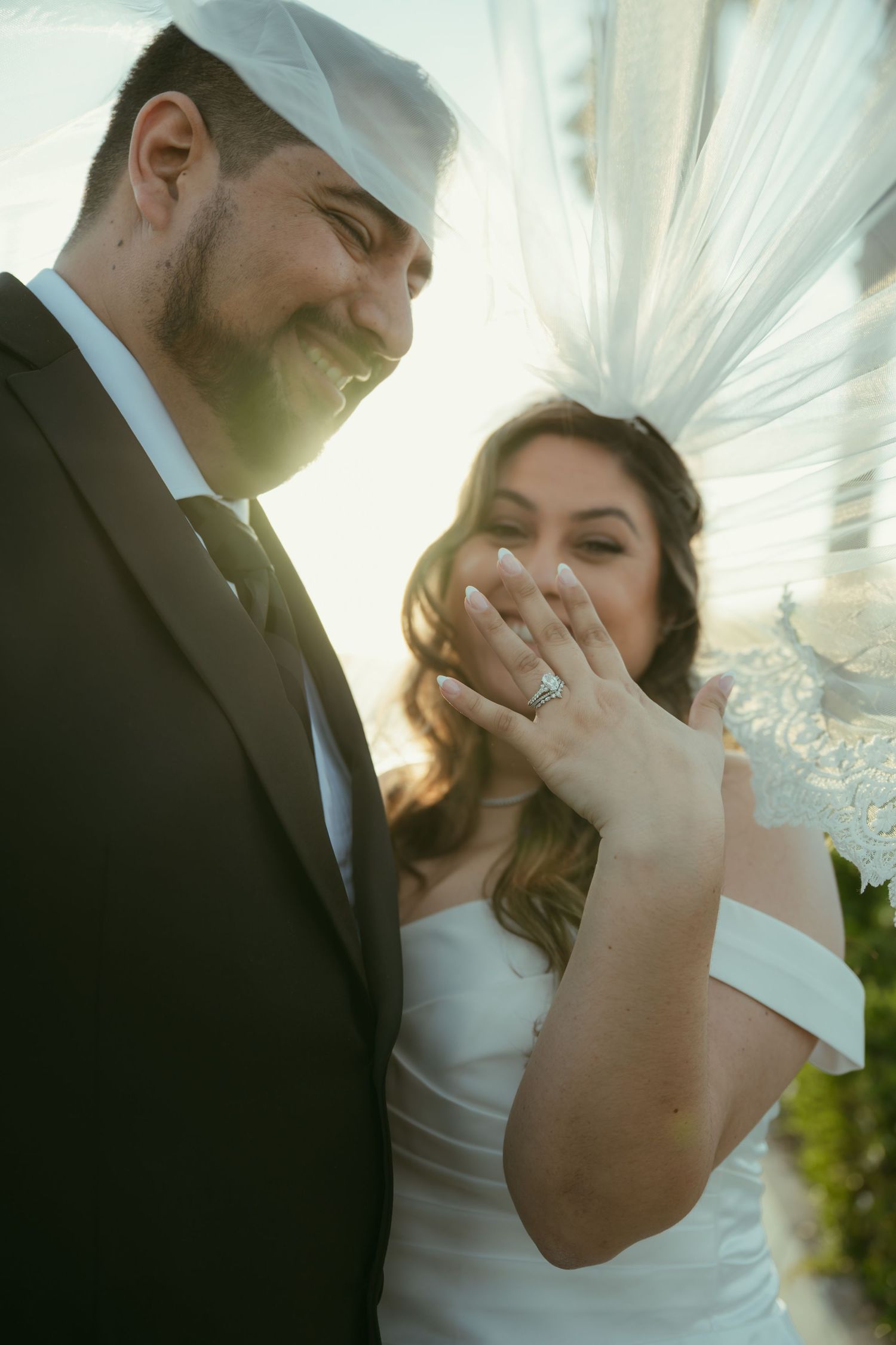 Smiling newlyweds pose under a veil as the bride shows off her wedding ring at a sunny outdoor ceremony.