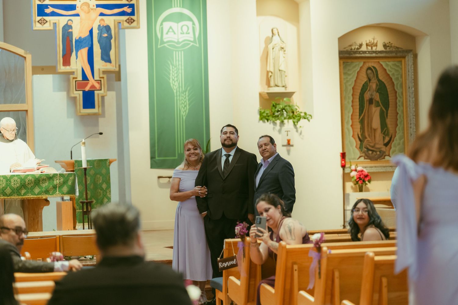 Couple stands together during a Catholic church wedding ceremony with guests seated in wooden pews and religious decor.