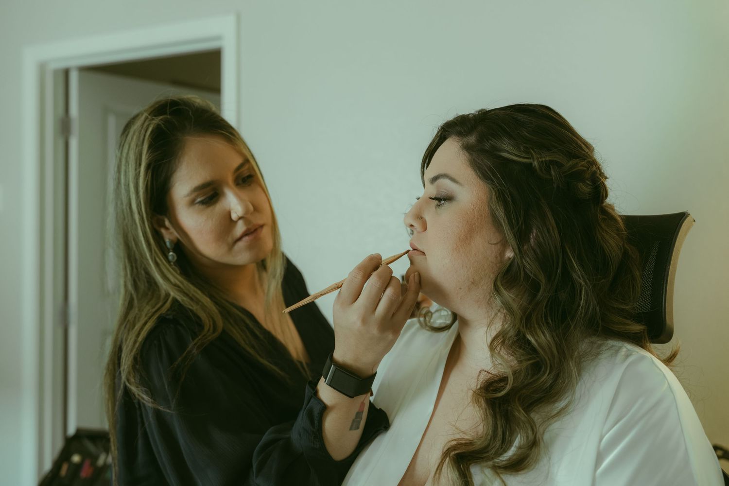 Makeup artist applying foundation to a bride in a white robe before her wedding day.