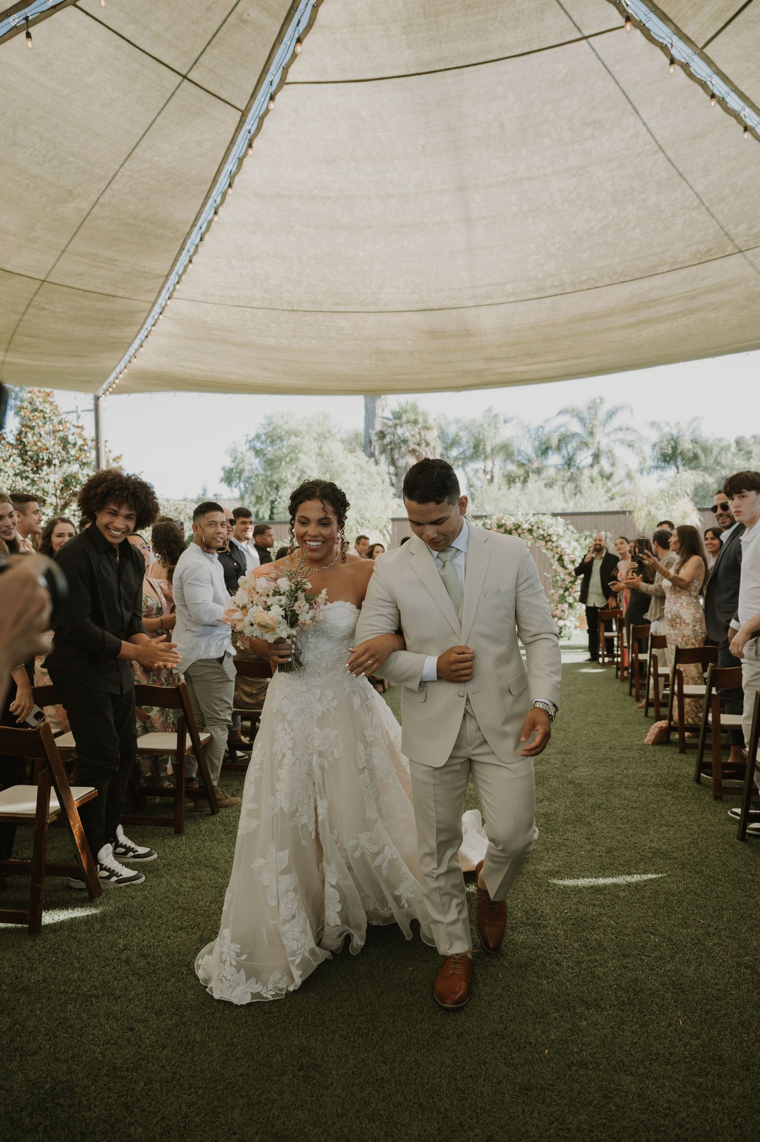Newlyweds exit ceremony aisle as bride holds bouquet in elegant lace gown beside groom in light suit.