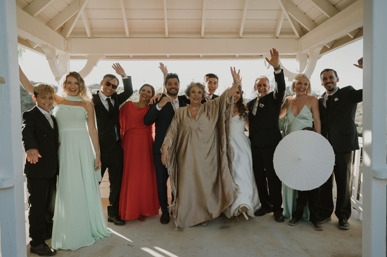 Large wedding party celebrating with raised arms under white gazebo at outdoor venue.
