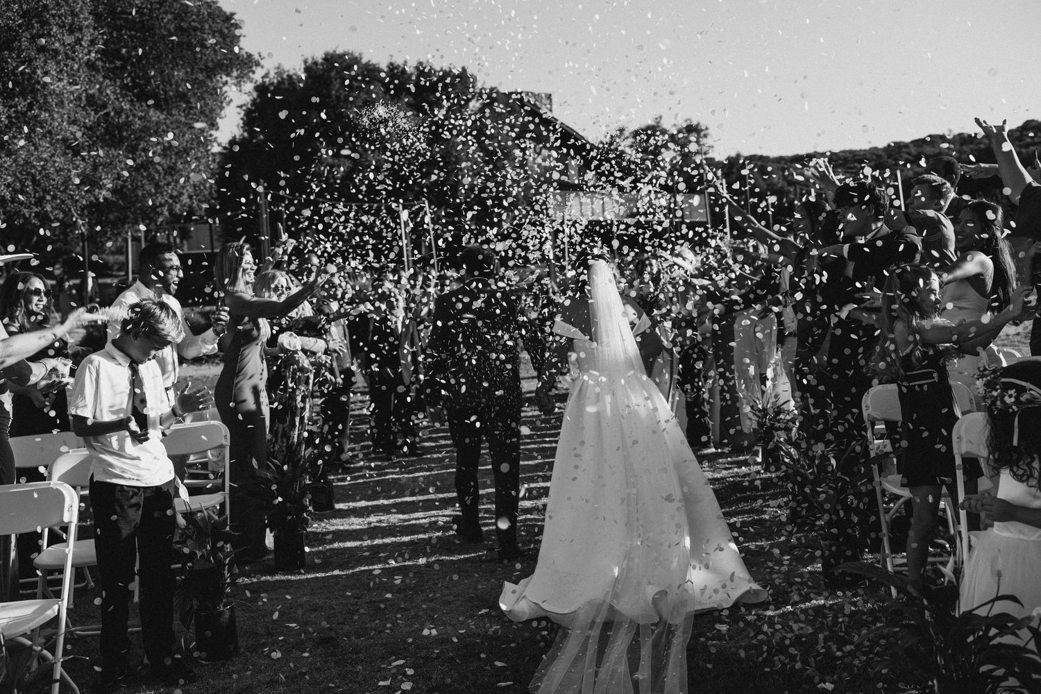 Black and white photo of bride and groom walking through confetti shower at outdoor wedding ceremony.