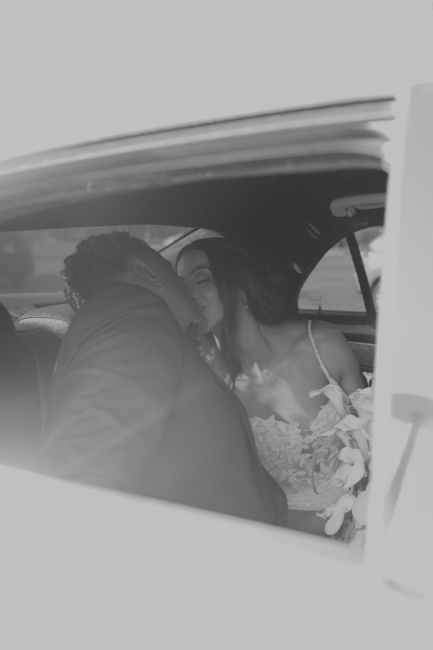 Bride and groom share a romantic moment in the backseat of a vintage car in black and white photo.