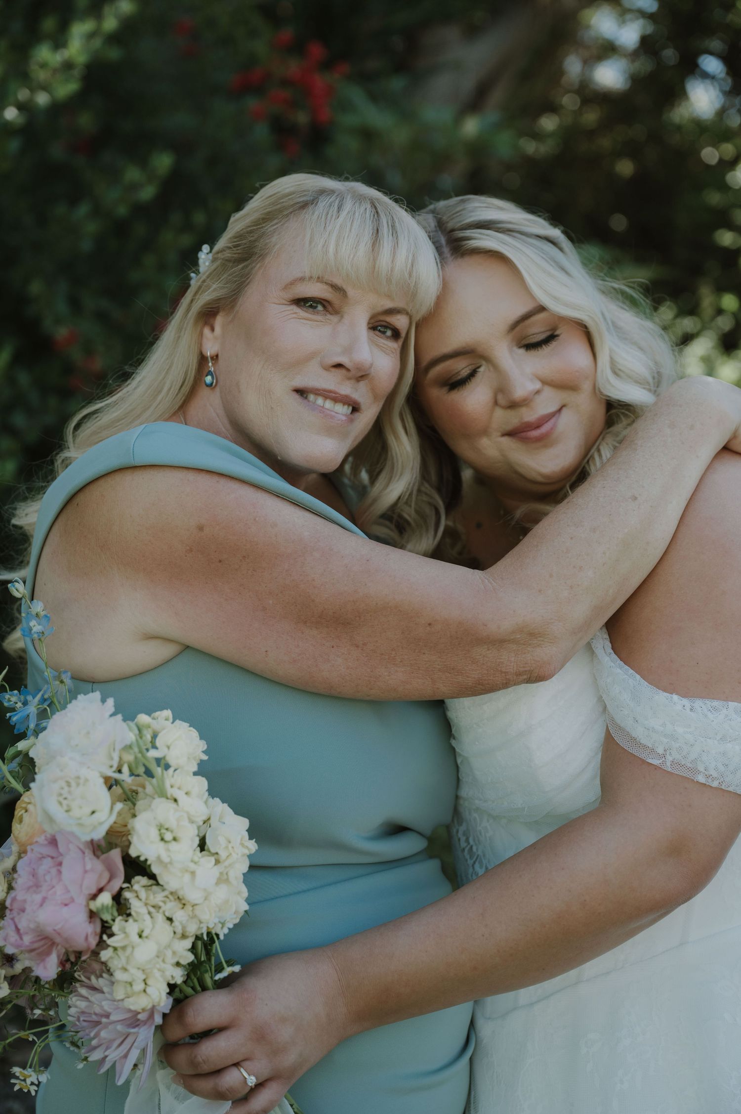 Bride in white gown embraces bridesmaid in sage dress holding floral bouquet outdoors.